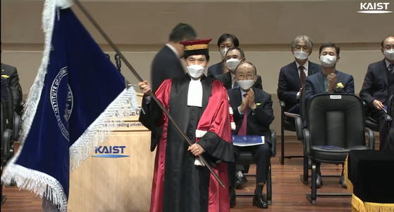 이광형 신임 KAIST 총장이 신성철 전 총장에게 교기를 인계받아 흔들고 있다. 뒷편으로 흰색 마스크를 착용한 김정주 NXC 대표(좌석 가운뎃줄 뒤에서 두 번째)가 웃고 있다. [유튜브 캡처]